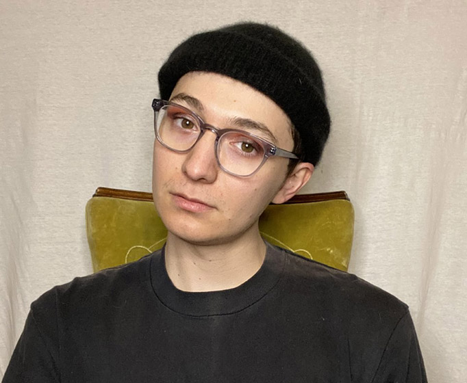 Person with head tilted to their left, wearing gray-rimmed glasses, a black beanie hat, and a black shirt.